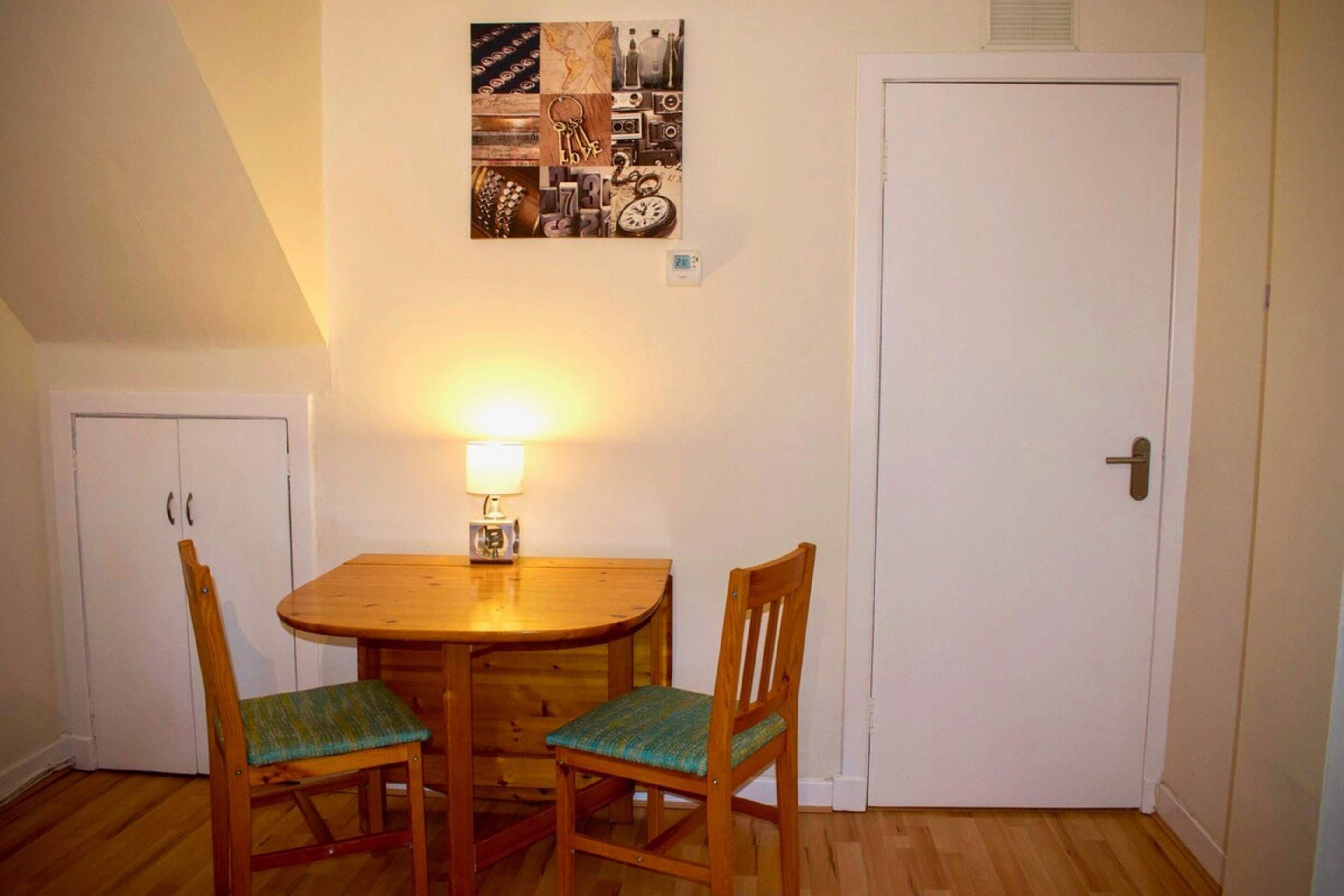 A dining area with a wooden table and two chairs, a wall clock, and a small table lamp.
