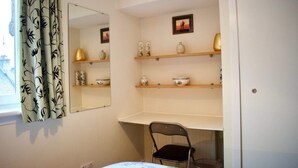 Ein Zimmer mit einem Bett, einem Sessel, einem Schreibtisch, Regalen mit Dekoration, einem Spiegel und einem Fenster mit Vorhängen.