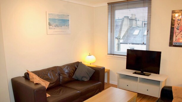 Ein Wohnzimmer mit einem braunen Ledersofa, einem Holztisch, einem Flachbildfernseher auf einem weißen Standfuß und einem Fenster mit Jalousien.
