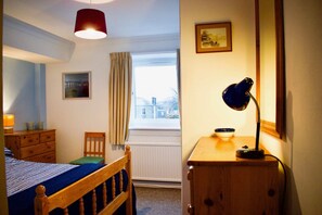 A bedroom with a bed, a wooden dresser, a desk with a lamp, a chair, a window with curtains, and a framed picture on the wall. - Welcoming and Homely 2 Bed in Central Location (Edinburgh)