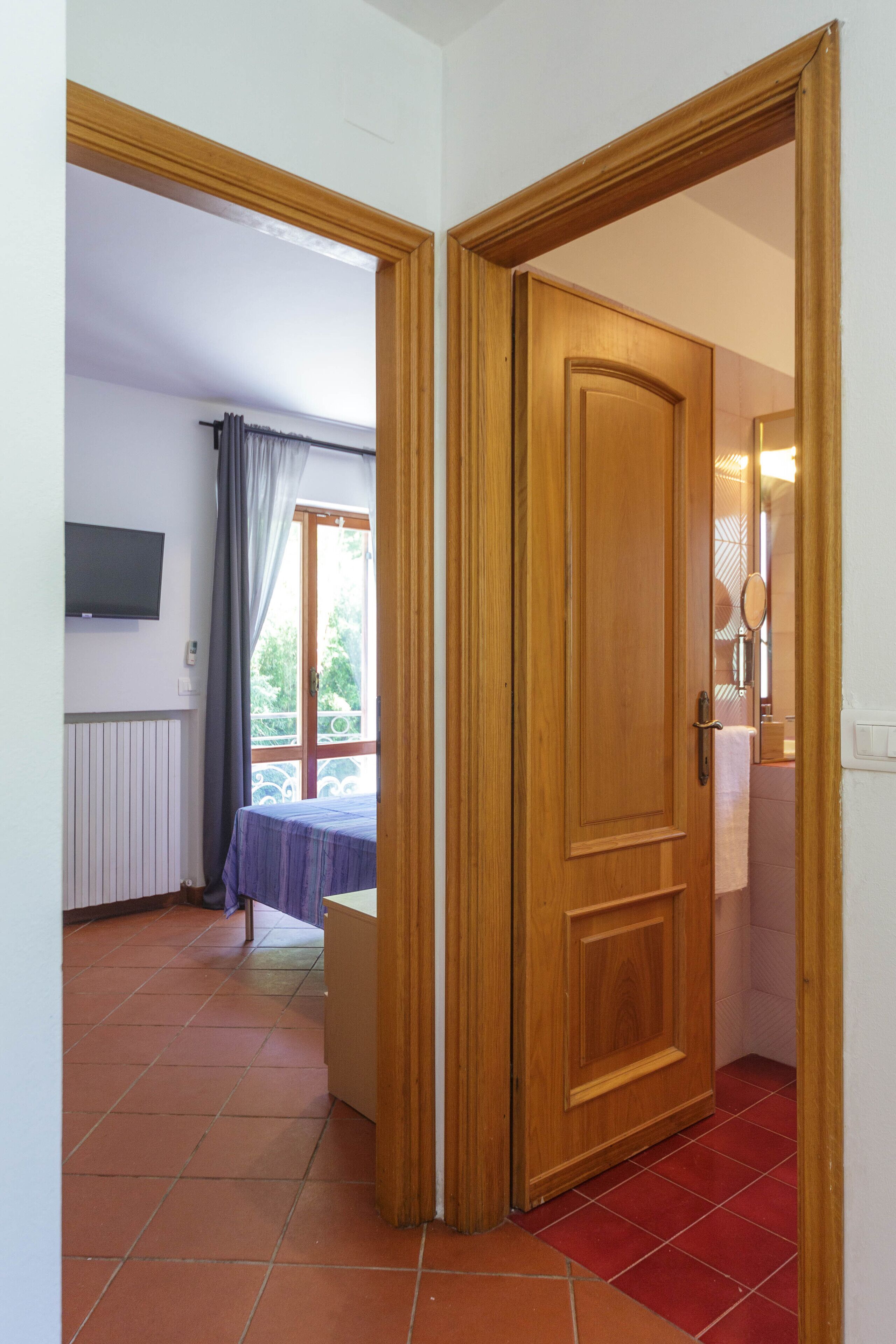 Quarto com porta de madeira, cama, televisão presa à parede e banheiro visível por uma porta aberta.