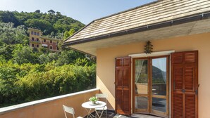 Uma varanda com mesa e cadeiras, uma porta de madeira e vista para uma colina com edifícios.