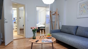 A room with a grey sofa, a round wooden coffee table with books and fruit, a vase with flowers, and a hanging coat rack. - Park 2- Charming 1-bdr Apt Close to Metro (Copenhagen)