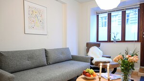 A living room with a grey sofa, a round wooden coffee table with candles and fruit, a white armchair, and a window with a view of buildings. - Park 2- Charming 1-bdr Apt Close to Metro (Copenhagen)