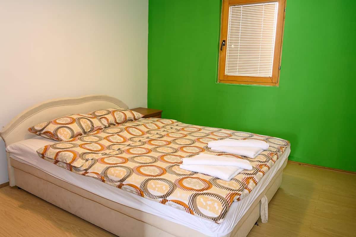 A bed with a patterned bedspread and pillows, a wooden nightstand, and a window with blinds.