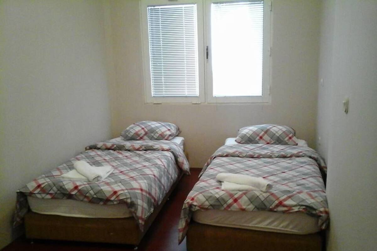 A small room with two single beds, each with a mattress and a blanket. There are two windows with blinds, and the walls are plain.