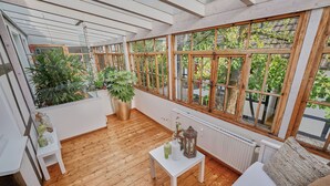 A room with wooden flooring, a white table with decorative items, large windows with wooden frames, and potted plants. - Apartment in the half-timbered courtyard 'house in motion' (Neunkirchen-Seelscheid)