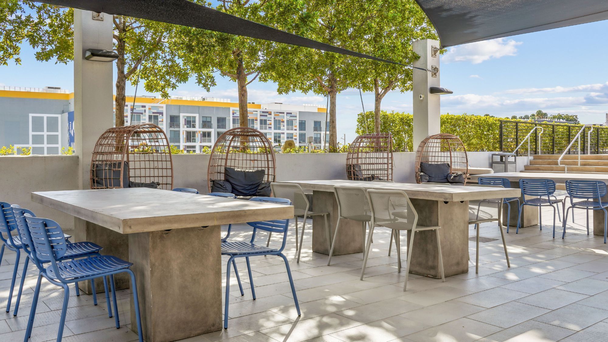An outdoor patio with concrete tables and blue chairs, surrounded by trees and a building in the background.