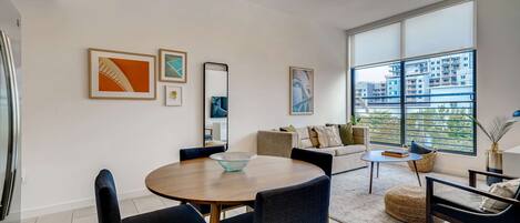 A modern living room with a round wooden dining table, dark blue upholstered chairs, a sofa, and a large window offering a view of buildings.