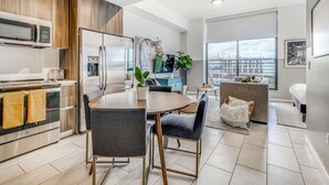 A modern kitchen with a dining area, a large window with a city view, and a living area with a sofa and a plant. - Sentral Wynwood (Miami)