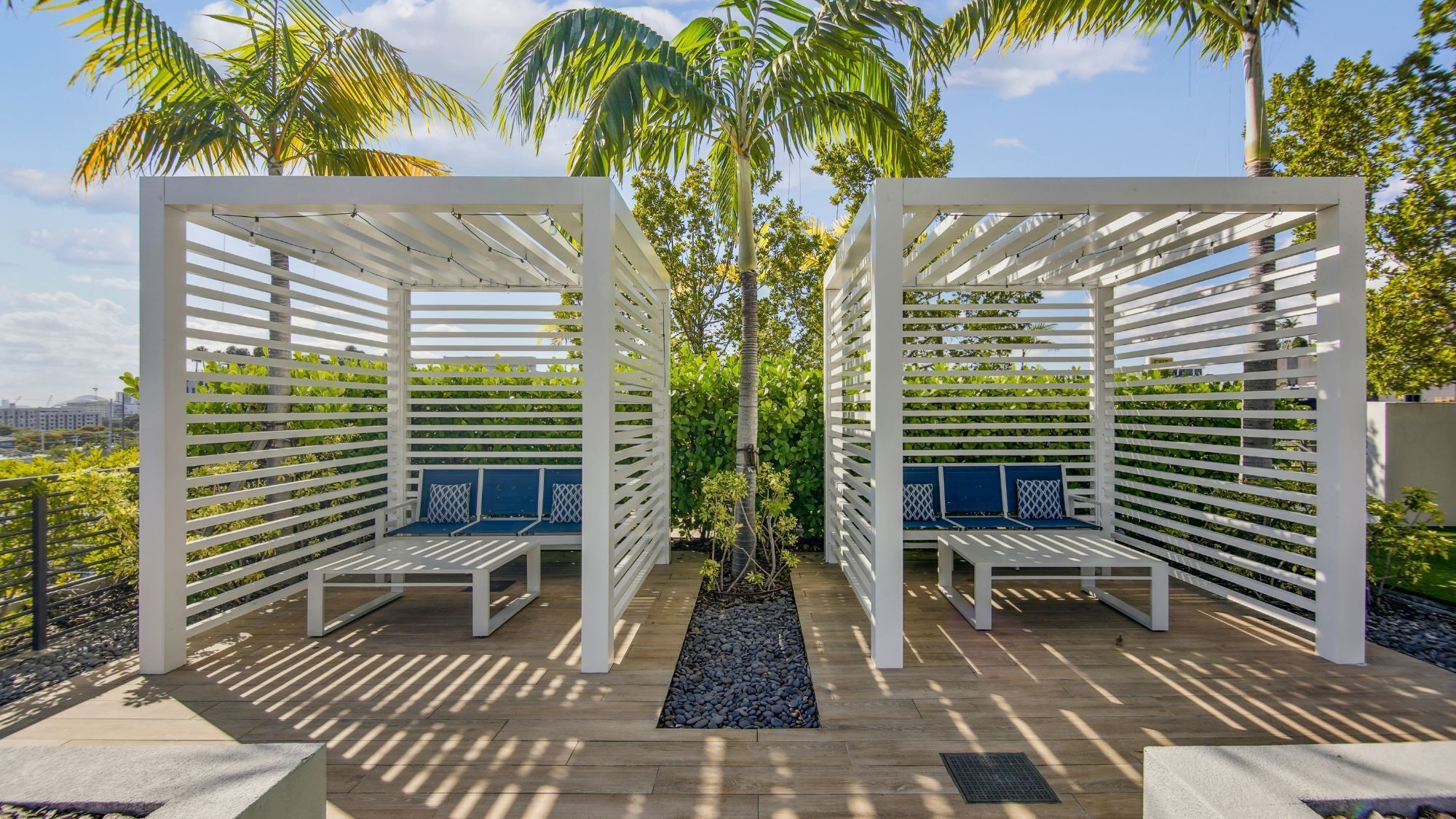 A covered outdoor seating area with white lattice structures and blue cushioned benches, surrounded by greenery and a view of the cityscape.