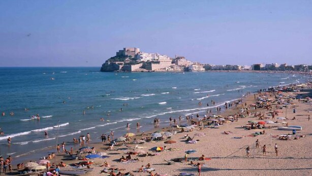Ein überfüllter Strand mit Menschen, die sich sonnen und schwimmen, ein großes Gebäude im Hintergrund und ein klarer, blauer Himmel.