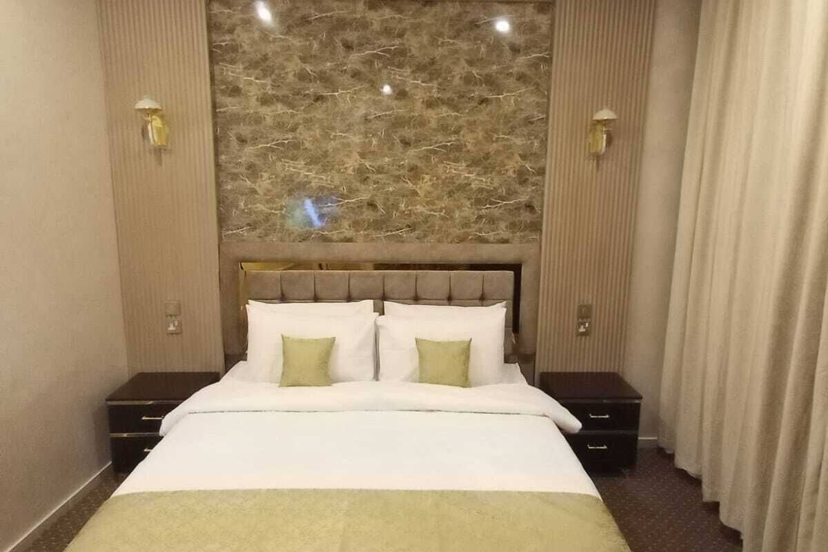 A hotel room with a large bed, two bedside tables, a headboard with a textured wall behind it, and a window with curtains.