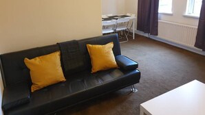 A living room with a black leather sofa, two mustard yellow cushions, a small table, and a window with curtains. - South Shield's Emerald 2 Bed Apartment (South Shields)
