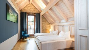 A bedroom with a wooden ceiling, a stone wall, a bed with white bedding, a blue chair, and a window with curtains. - Ferienwohnungen Alte Destillerie Rathen (Rathen)
