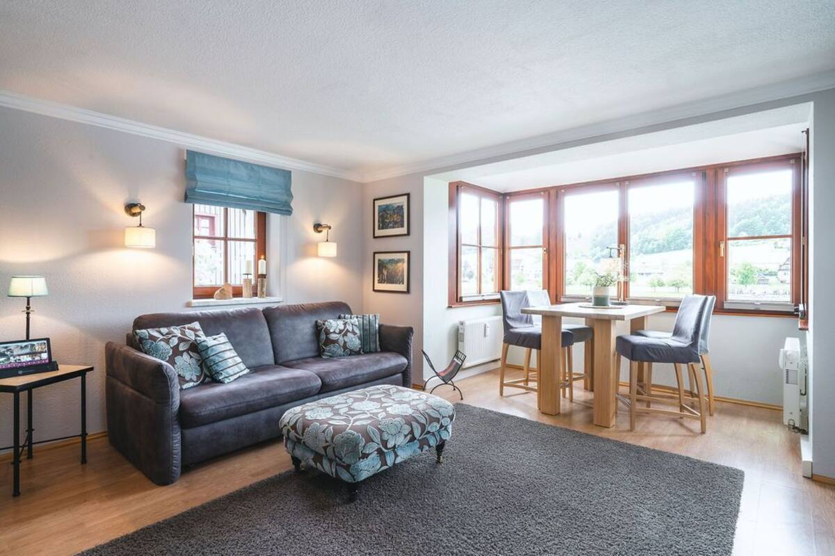 Ein Wohnzimmer mit Sofa, Hocker und Esstisch mit Stühlen. Es gibt Fenster mit Vorhängen, Wandlampen und gerahmte Bilder.