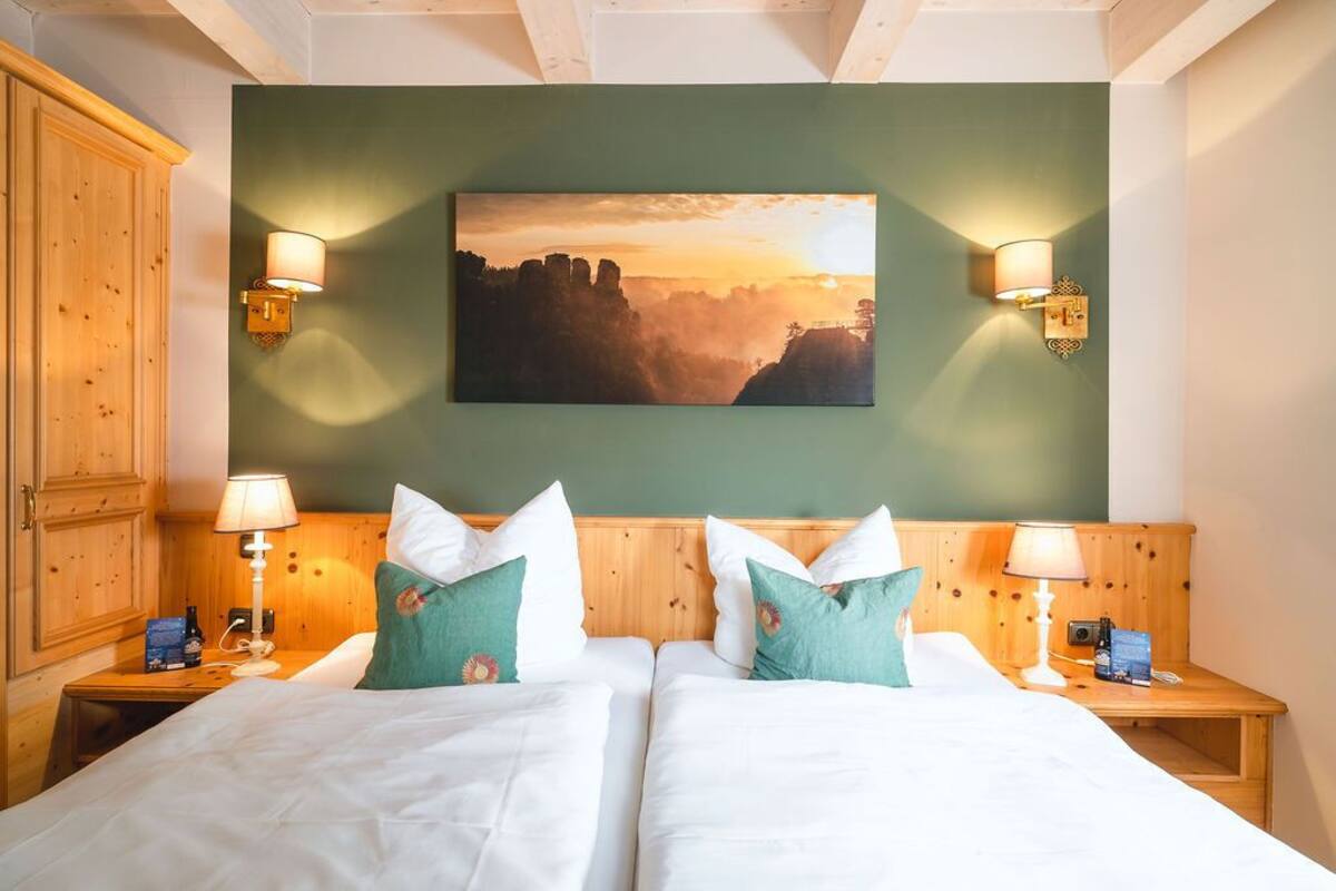 Ein Doppelbett mit weißer Bettwäsche, zwei Kissen, zwei Nachttische mit Lampen und ein Gemälde mit Sonnenuntergang über einer bewaldeten Landschaft an der Wand.