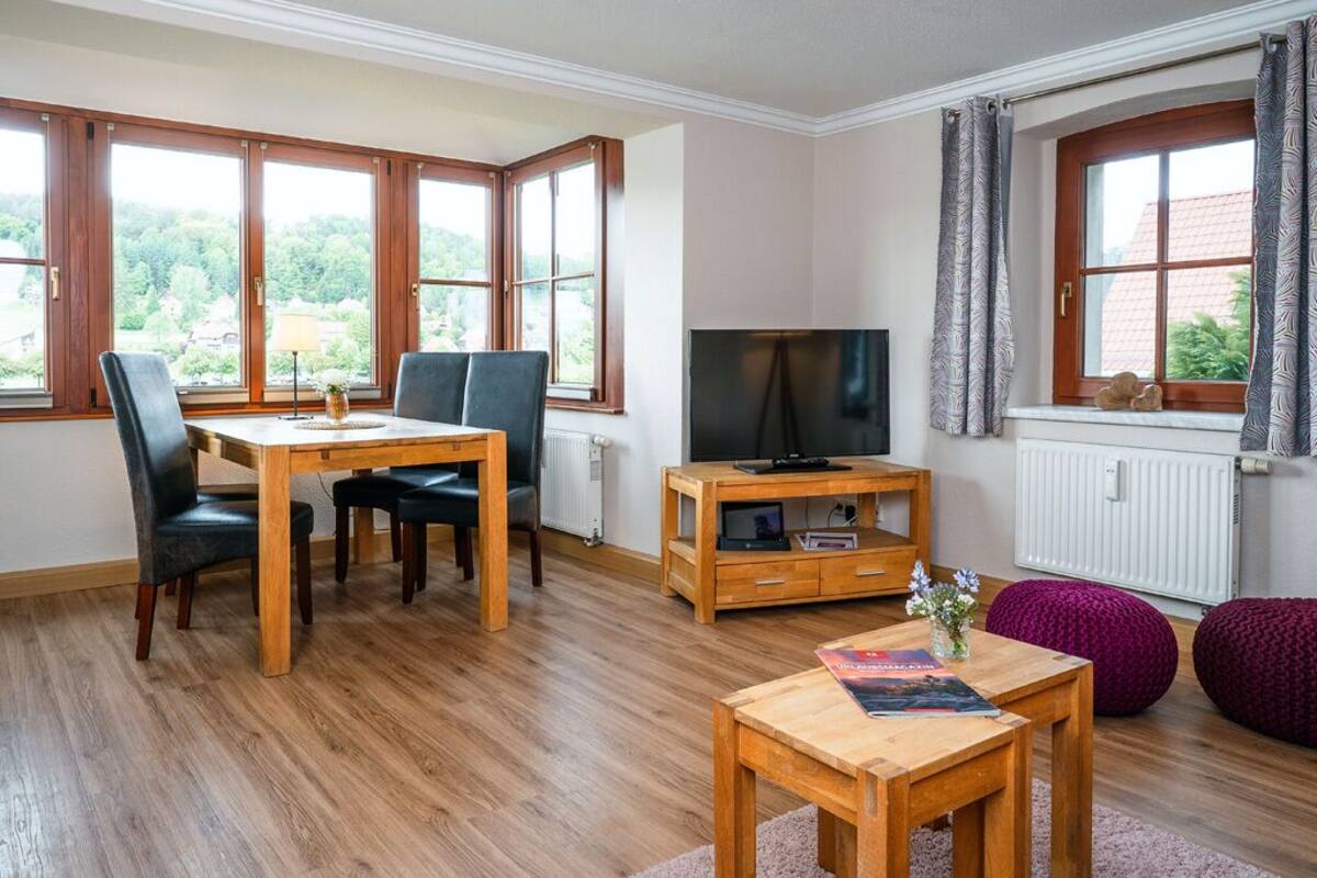 Ein gemütliches Wohnzimmer mit einem Holzesstisch, zwei Stühlen, einem Fernseher auf einem Holzschrank und einem Fenster mit Vorhängen.
