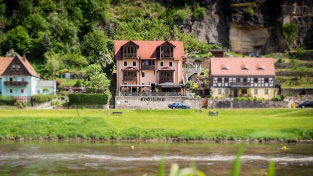 A building labeled "DESTILLERIE" with a red roof, situated by a river with a grassy area in front. - Ferienwohnungen Alte Destillerie Rathen (Rathen)
