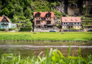 Un bâtiment portant l’inscription « DISTILLERIE » et surmonté d’un toit rouge, situé au bord d’une rivière, avec une zone herbeuse devant.
