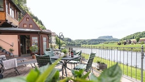 An outdoor patio with a table and chairs, overlooking a river and a hill. - Ferienwohnungen Alte Destillerie Rathen (Rathen)
