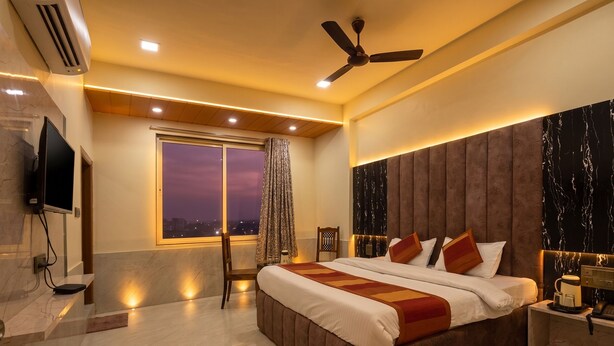 A modern hotel room with a large bed, a ceiling fan, a TV, and a view of the city at dusk. - Hotel Star Palace (Jaipur)
