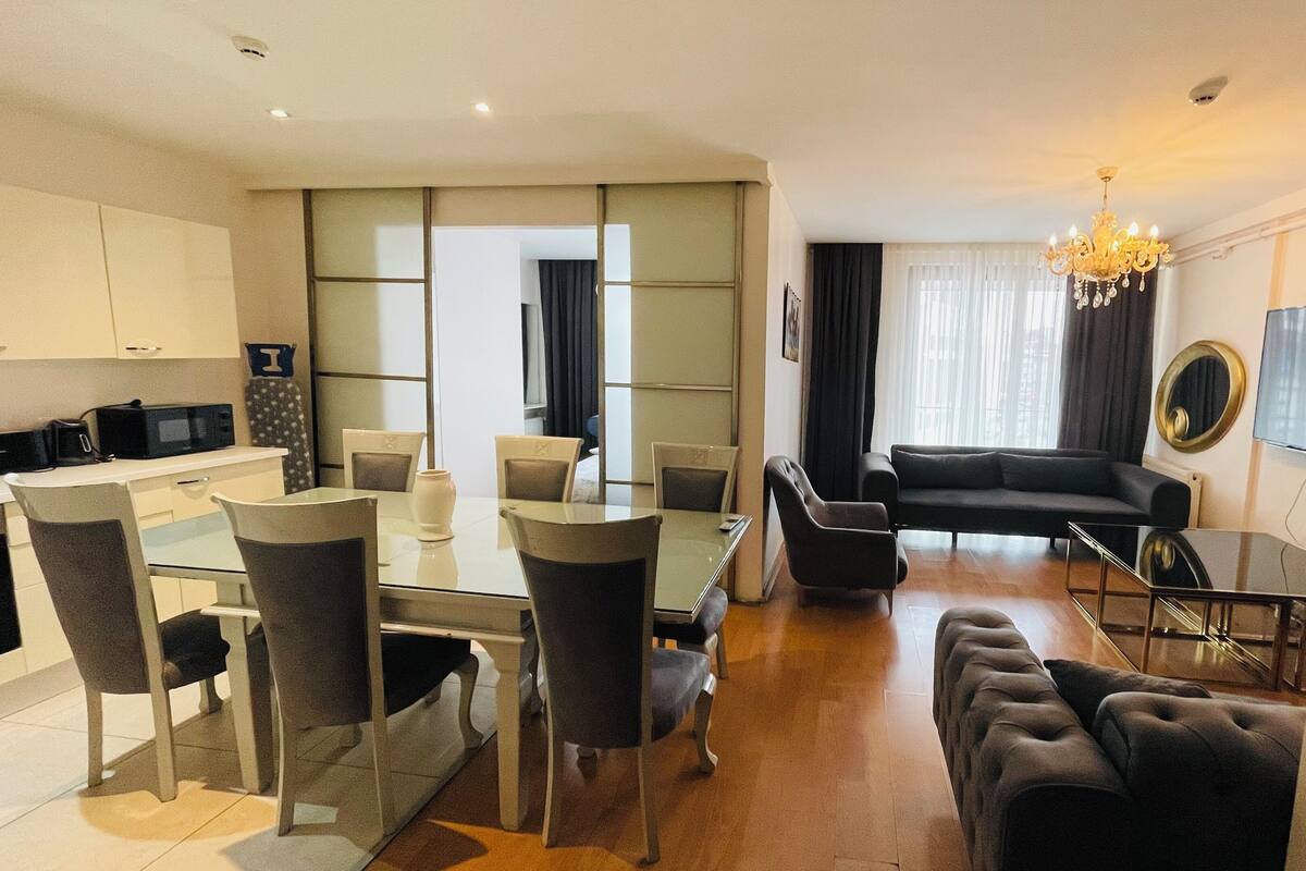 A modern hotel room with a dining area, a living area with a sofa and a TV, and a kitchen with a microwave.