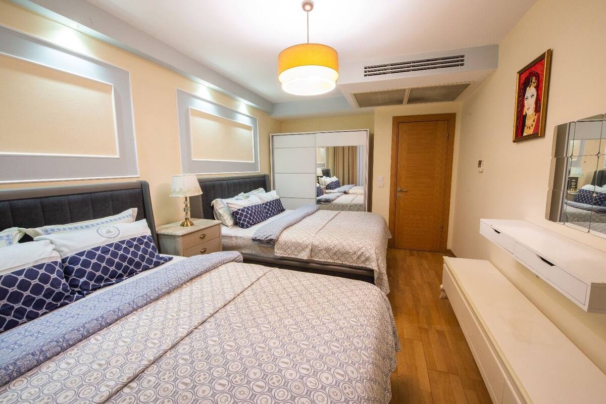 A hotel room with two beds, a bedside table with a lamp, a mirror, a wardrobe, and a small window.