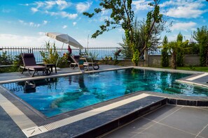 Une piscine entourée de verdure et d’une clôture, avec des chaises longues et un parasol.
