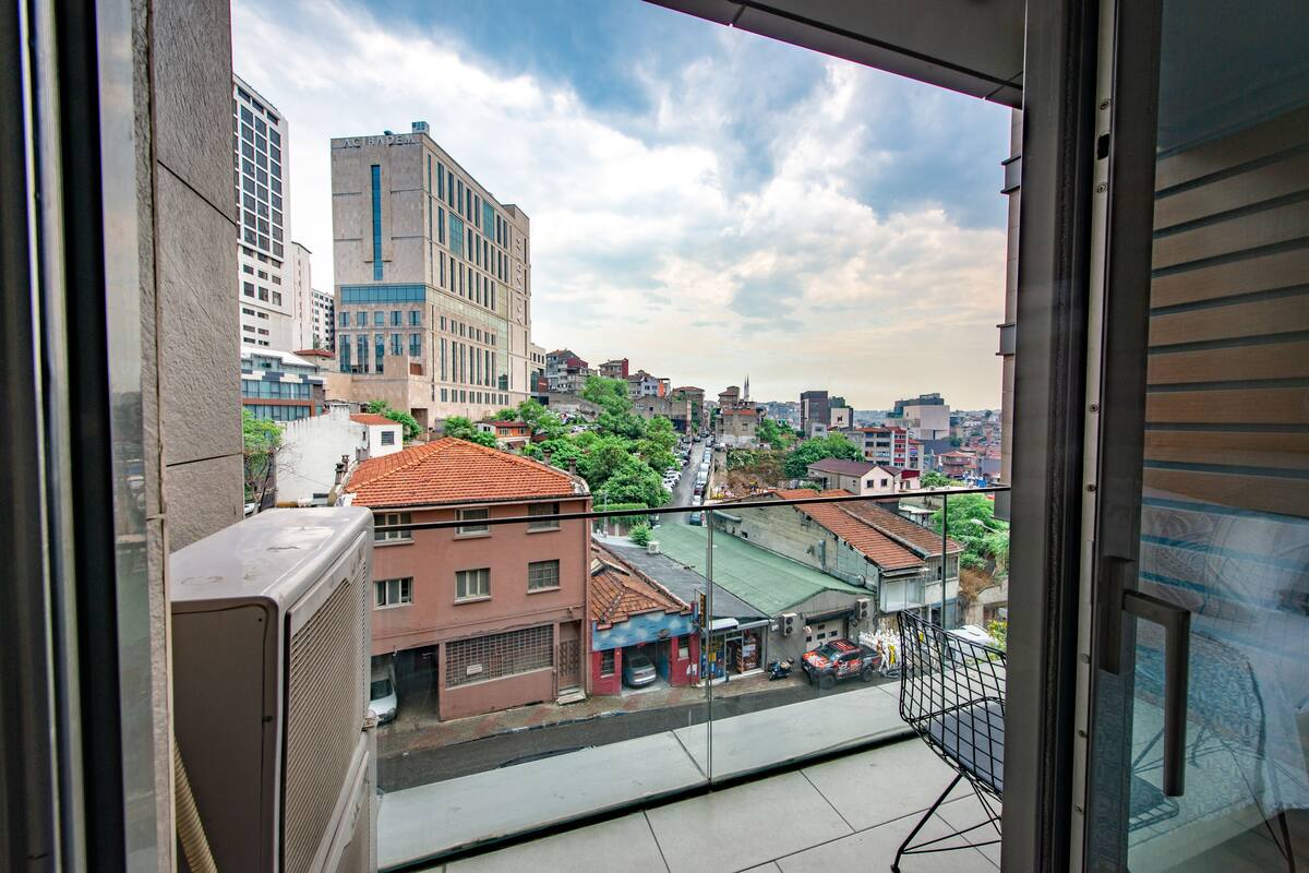 A view from a balcony overlooking a cityscape with buildings, a street, and a green area.
