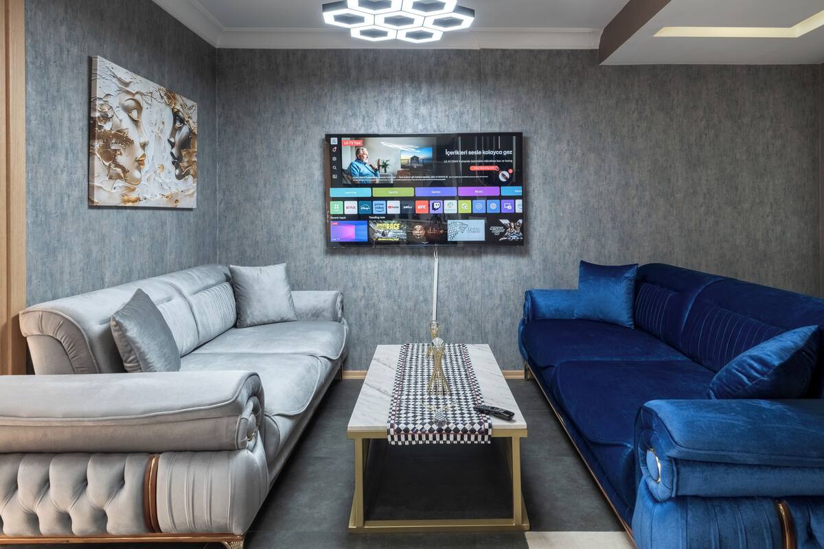 A modern living room with a grey sofa, a blue sofa, and a coffee table with a checkered table runner. A flat-screen TV is mounted on the wall displaying various channels.