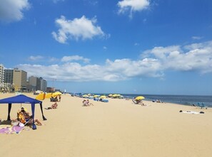 Una playa con tiendas de campaña y sombrillas, cielo despejado y edificios al fondo.