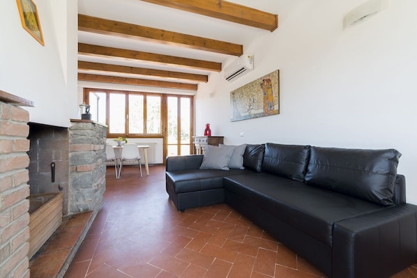 A living room with a black leather sofa, a stone fireplace, wooden beams, a tiled floor, and a window with natural light.