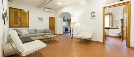A spacious living area with a sofa, a white armchair, and a floor lamp. There is a wooden cabinet and a door leading to another room.
