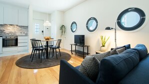 A modern kitchen with a built-in oven, a dining area with a round table and chairs, a TV on a stand, and a sofa with patterned cushions.