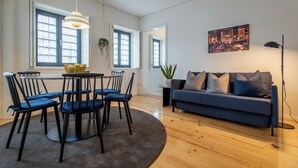 A modern living room with a blue sofa, a dining table with chairs, and a large window with curtains.