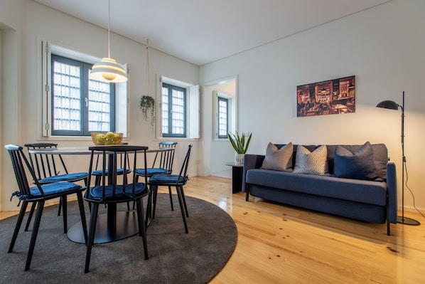 A modern living room with a blue sofa, a dining table with chairs, and a large window with curtains.