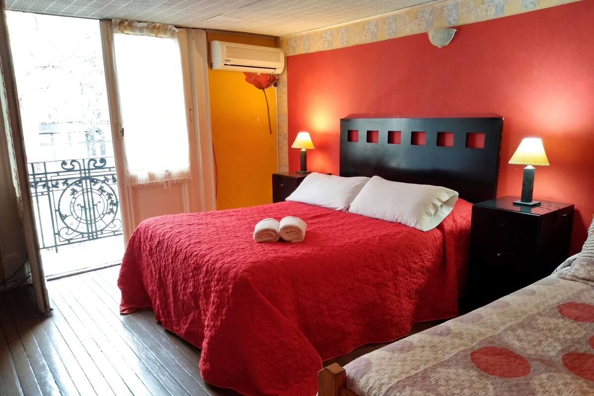 A hotel room with a red bed, two bedside lamps, a wooden headboard, and a balcony with a railing.