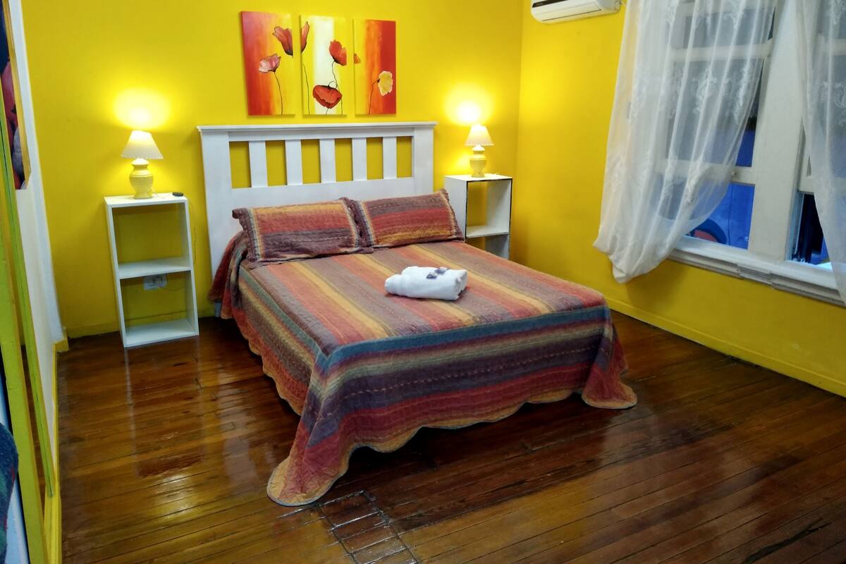 A room with a bed, two bedside lamps, a white headboard, a window with sheer curtains, and a wall-mounted air conditioner.