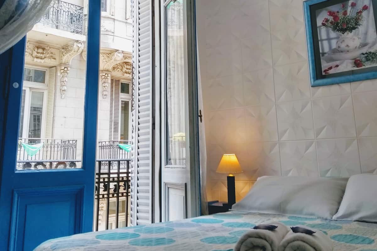 A hotel room with a bed, a nightstand, a lamp, a window with a view of a building, and a framed picture on the wall.