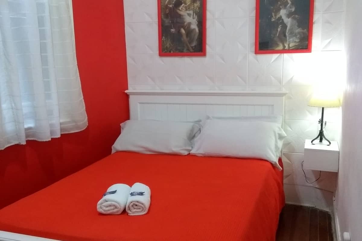 A hotel room with a red bedspread, white headboard, two framed pictures on the wall, a bedside lamp, and a window with sheer curtains.
