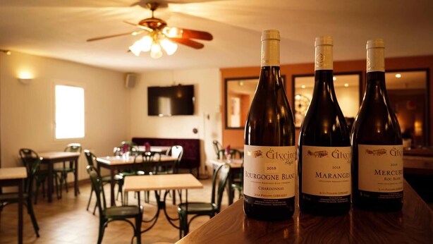 Three bottles of wine on a wooden table in a dining area with chairs and tables, a ceiling fan, and a TV on the wall. - Hôtel Restaurant - Les Jardins du Lac (Autun)