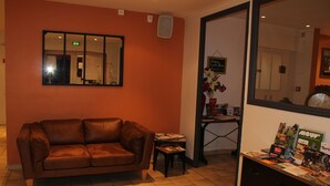 A room with a brown leather sofa, a small table with magazines, a mirror on the wall, and a door leading to another room. - Hôtel Restaurant - Les Jardins du Lac (Autun)