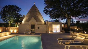 A pool area with lounge chairs, a stone wall, and a building with a conical roof. - TD Trullo Roverella Charming Trullo with Pool (Castellana grotte)