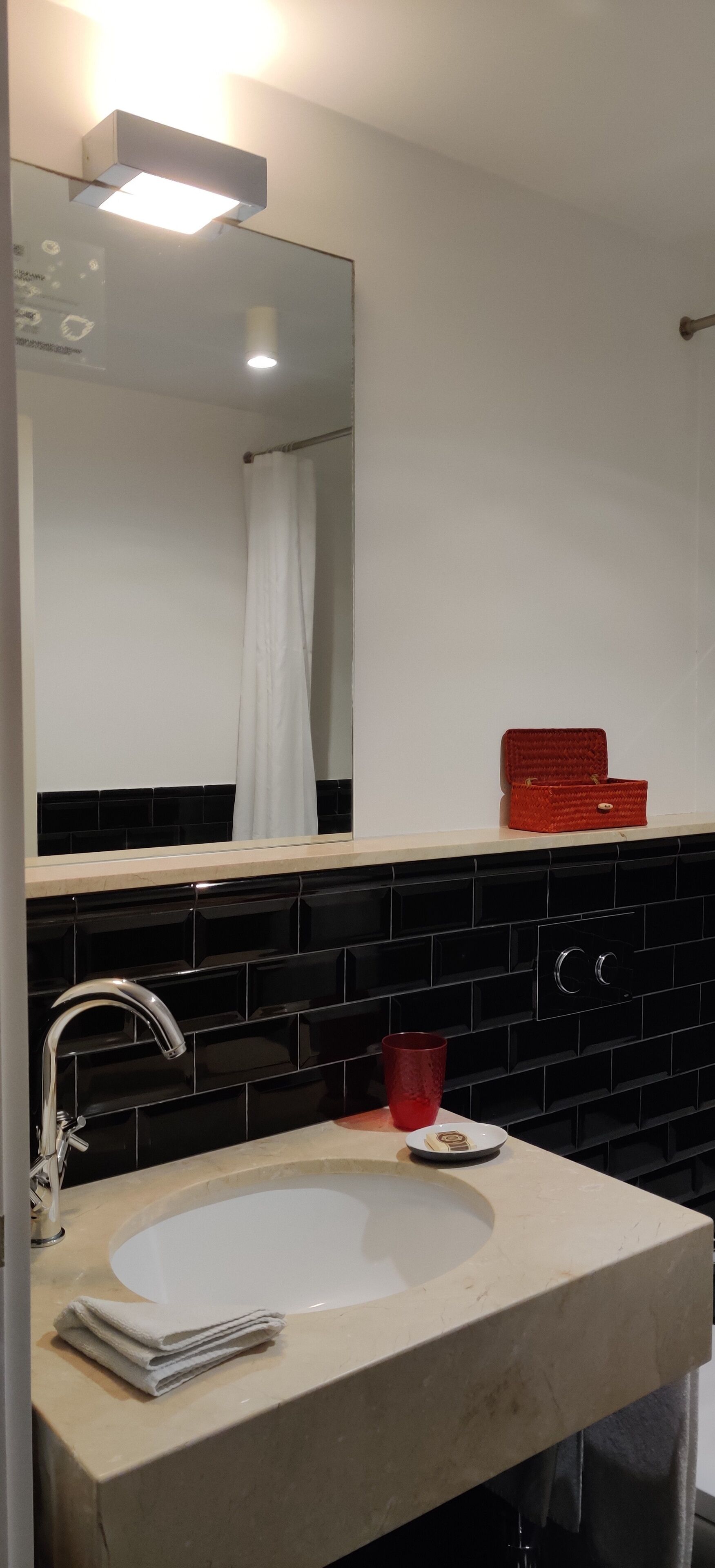 Un baño con lavamanos blanco, salpicadero de azulejos negros, una caja roja sobre la encimera y un espejo por encima del lavamanos.