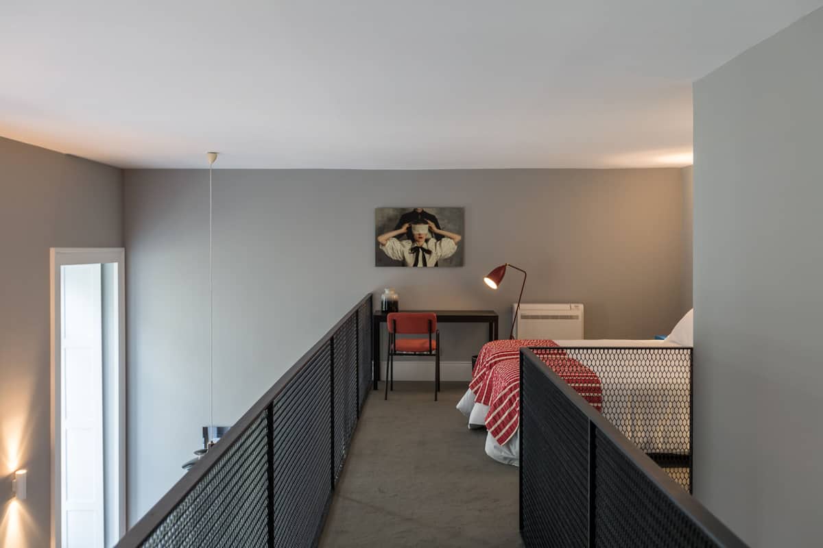 A compact bedroom with a bed, a desk, and a chair. There is a painting on the wall and a lamp on the desk.