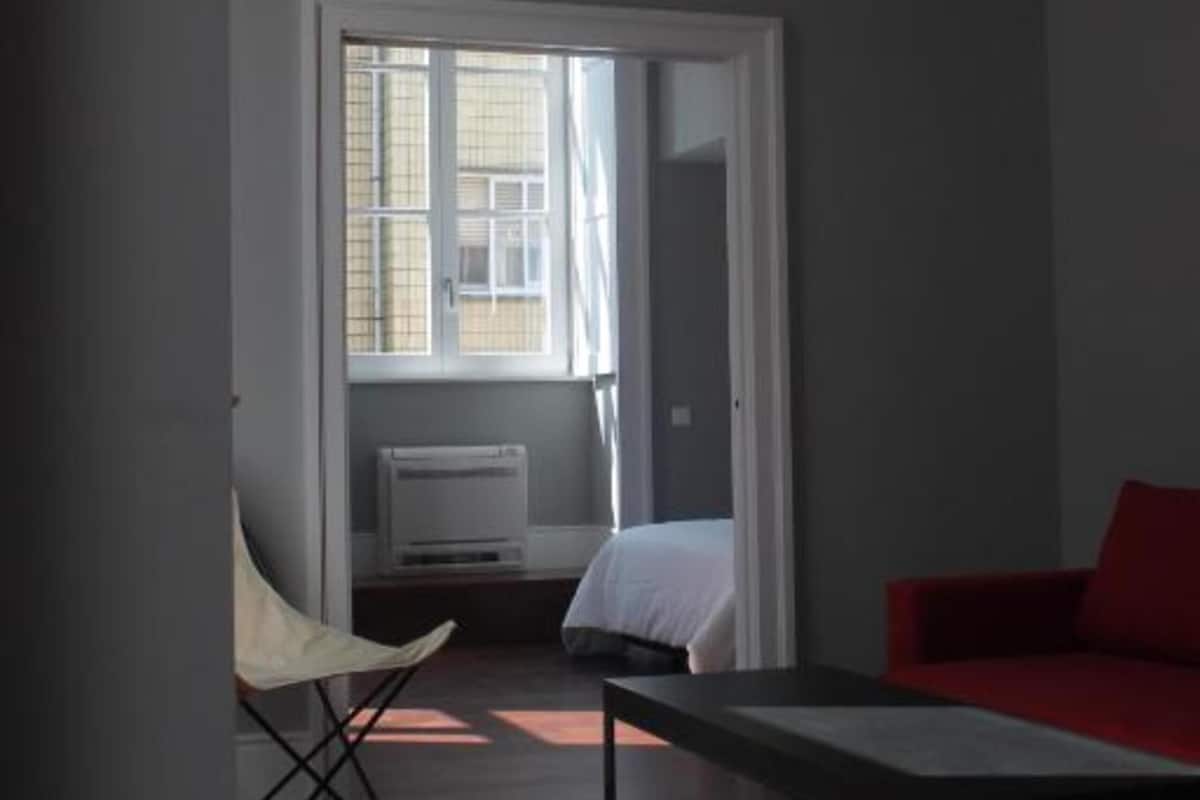 A room with a bed, a red sofa, a chair, and a window with sunlight casting shadows on the floor.