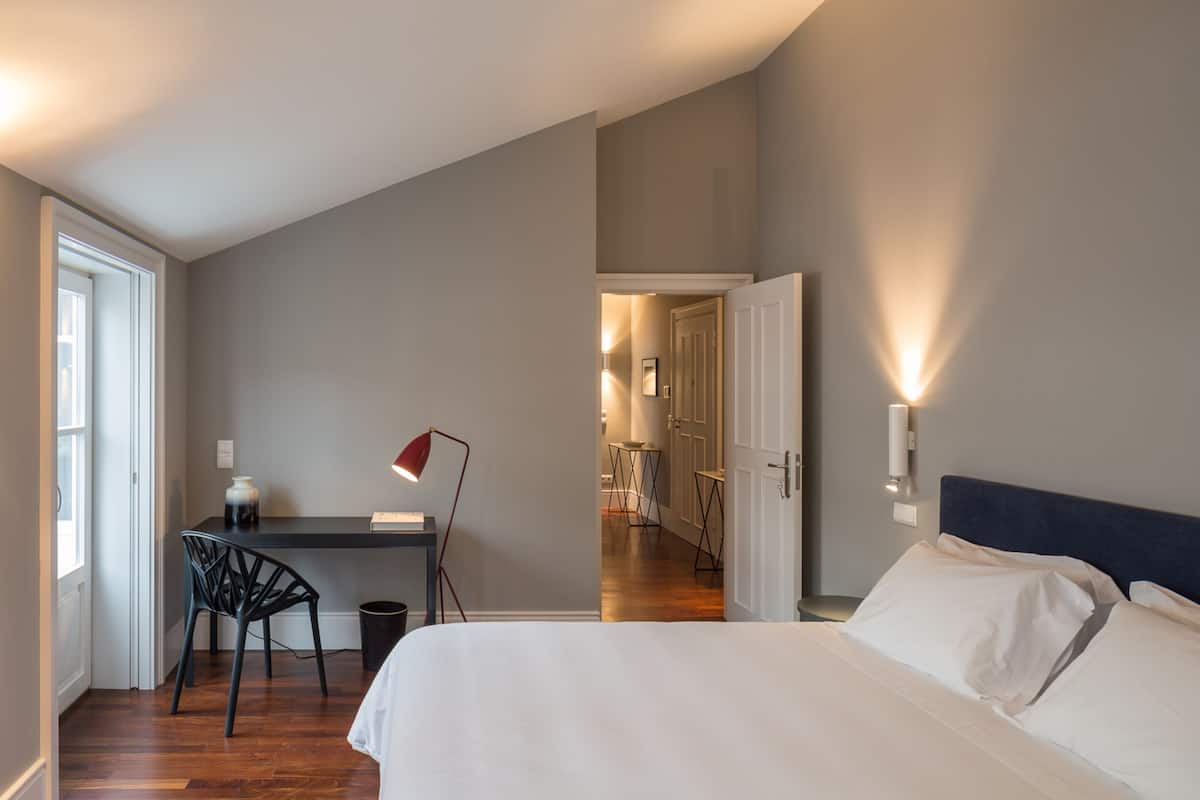 A hotel room with a bed, a desk with a chair, a lamp, and a view of the hallway.