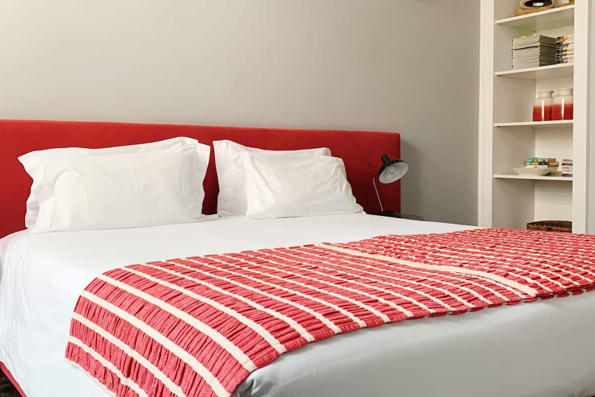 A bed with white bedding and a red headboard, a bedside table with a lamp, and a white shelving unit with various items.