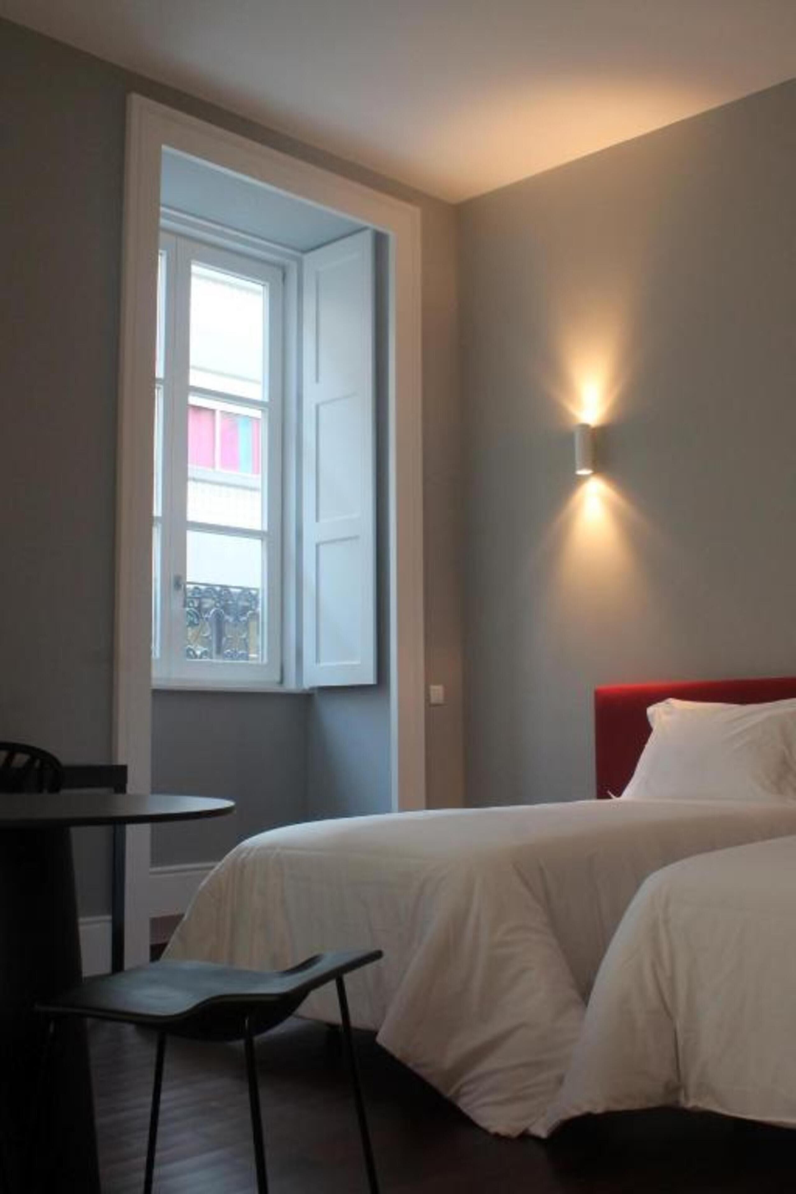 A bedroom with a bed, a chair, a table, a window, and a wall-mounted light.